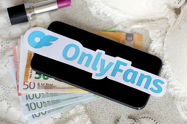  Evro bankovci in mobilni telefon čez njiju je napis OnlyFans, poleg leži ženska šminka za ustnice.